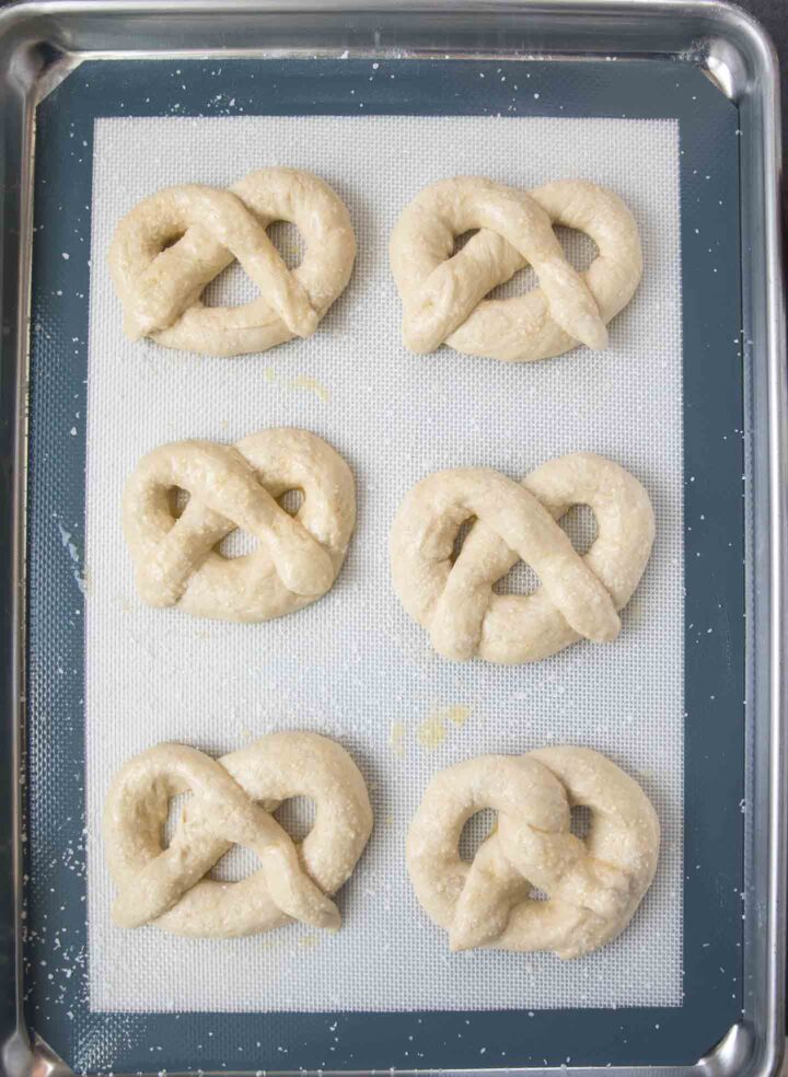Six homemade soft pretzels shaped pieces of dough unbaked on a cookie sheet topped with an egg wash.
