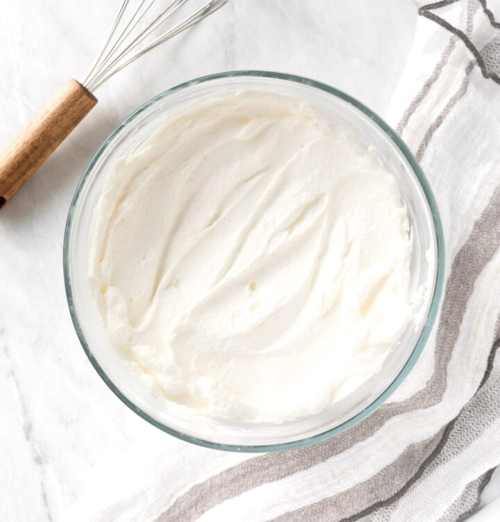 Homemade whipped cream in a clear bowl with a napkin and whisk.