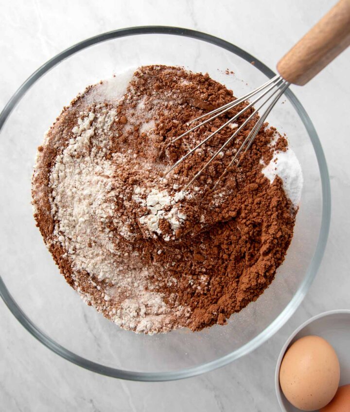 Dry ingredients of four, sugar, salt, baking soda, baking powder, unsweetened cocoa in a bowl with a whisk.