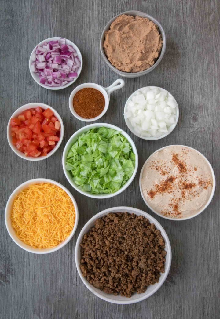 7 layer dip ingredients with purple onions, taco seasoning, diced white onions, diced tomatoes, shredded lettuce, shredded cheese, refied beans and seasoned taco meat in individual small white bowls on a gray wood background.