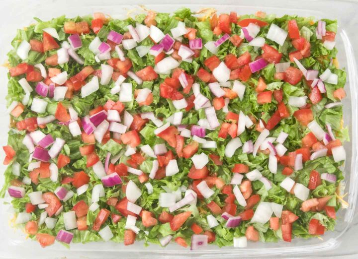 7 Layer Dip with shredded lettuce, diced tomato, red and white diced onion in a 9x13 baking dish.