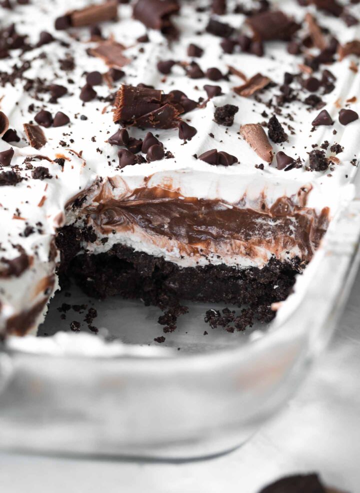 Chocolate Lasagna in a baking dish with a slice taken out of it. Showing the layers of Oreo crust, cream cheese, chocolate pudding and Cool Whip topping. Garnished with mini chocolate chips and chocolate shavings.