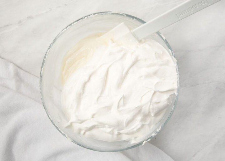 A clear bowl with whipped cream cheese, sugar and Whip Cream and a white spatula.